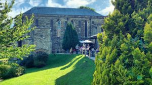 A green lawn in the sunshine. On the right of the image people sit at tables with white umbrellas.