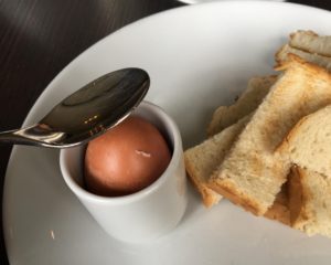 A spoon cracks the top of a boiled egg. It is on a white plate with lots of toast.
