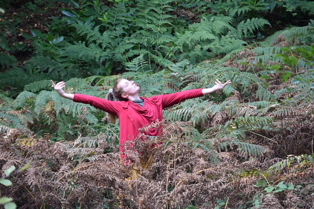 A young dancer is standing amongst ferns and bracken, holding her arms out to the side and looking to the sky. She is wearing a loose, dark red long sleeved top, and has brown hair tied back in a ponytail.