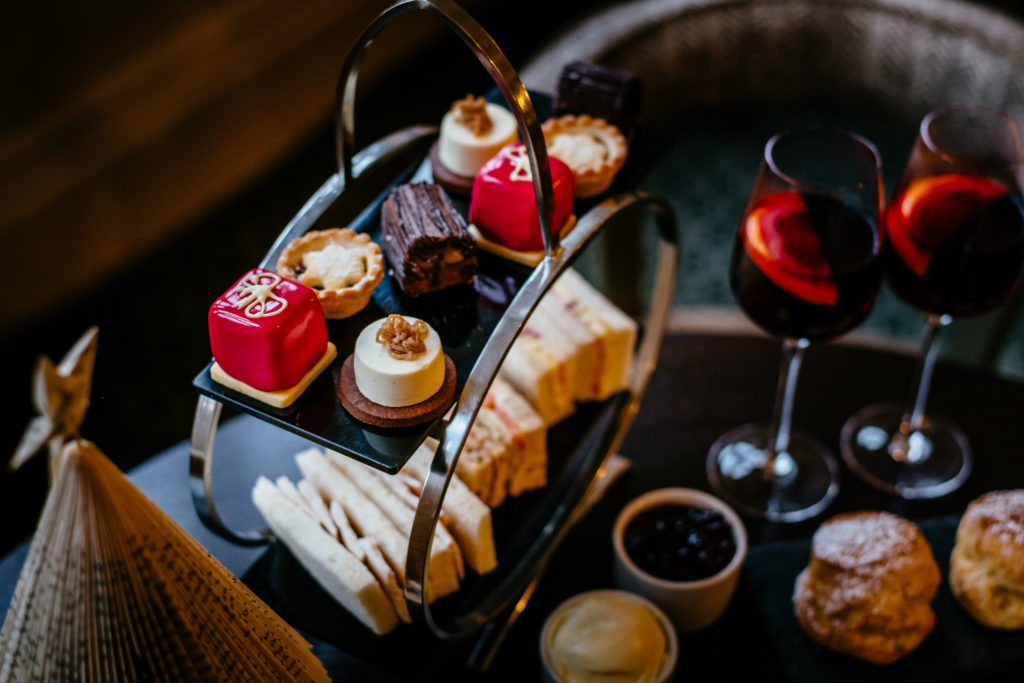 A table is set with a large circular stand which holds neat, small sandwiches, and perfectly tiny cakes and pastries for afternoon tea. On the table next to it are two glasses of mulled wine, a pot of jam, a pot of clotted cream, and two icing sugar dusted scones.