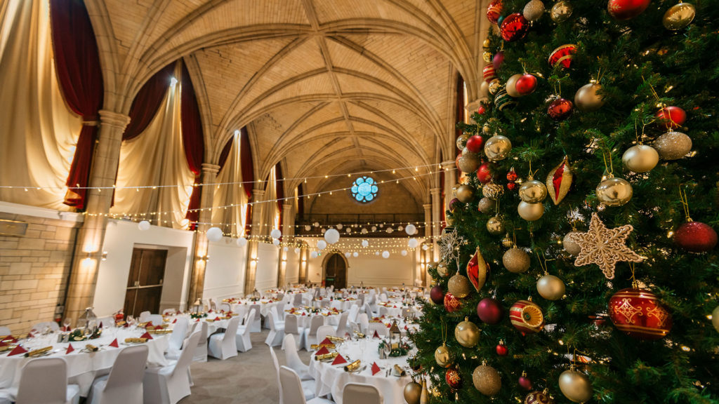 The interior of Alverton Hotel in Truro. The image shows the great hall, with a high arched ceiling and white covered chairs set out around white covered tables. Fairy lights are strung across the room, and a large Christmas tree is in the foreground of the photo. It is decorated with red and gold baubles.