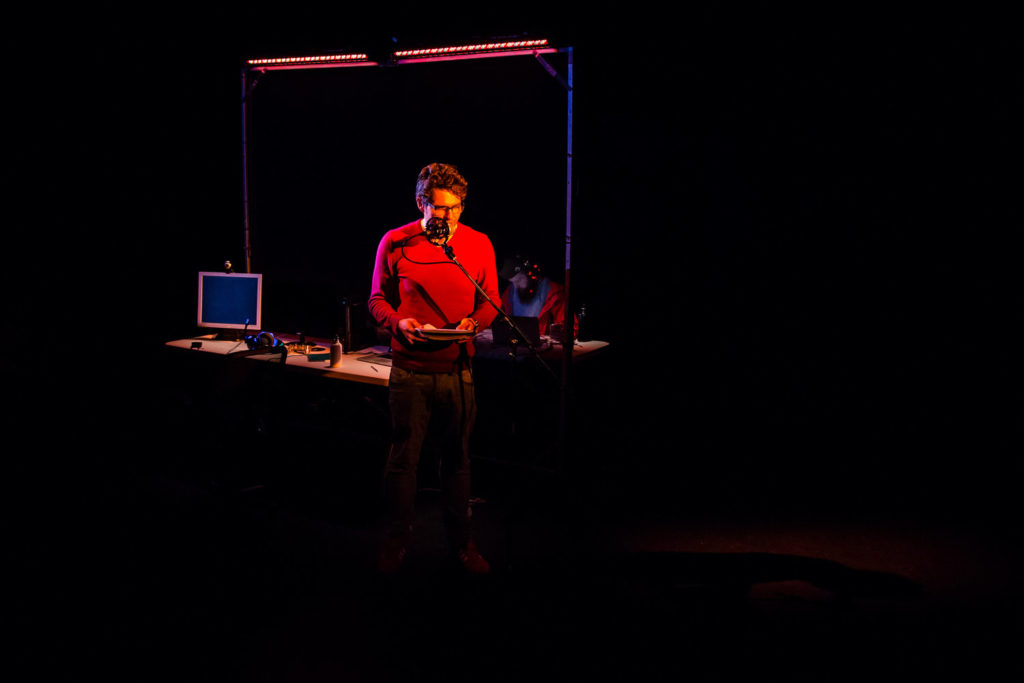 A man in a red shirt stands on stage, with a black background behind him
