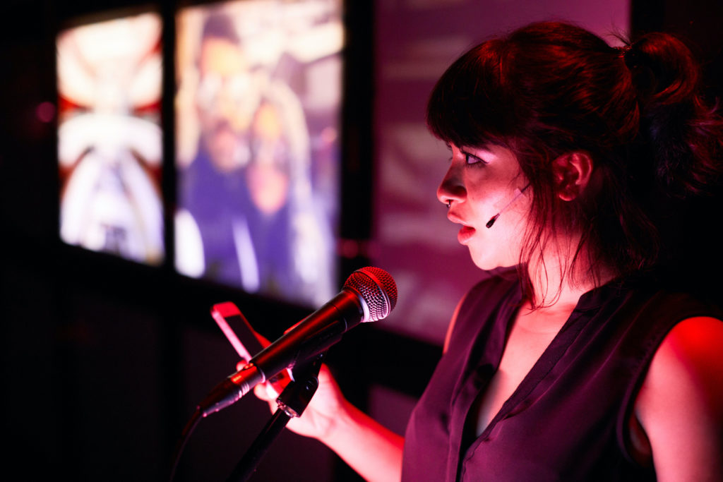 A woman with dark brown hair is standing with a phone in her hand, speaking into a microphone.