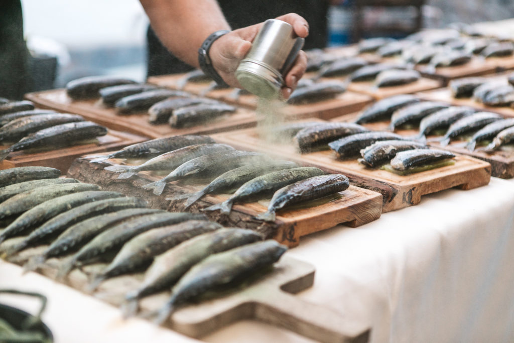 Someone dusts whole fish with some kind seasoning. The fish are lined up on wooden boards.