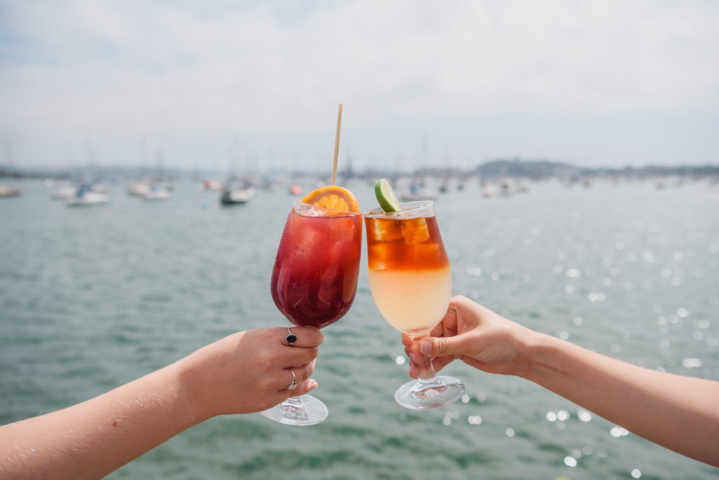 Two people, off camera, are clinking two orange cocktails together. We see their arms and hands, and the blue glittering sea behind them.