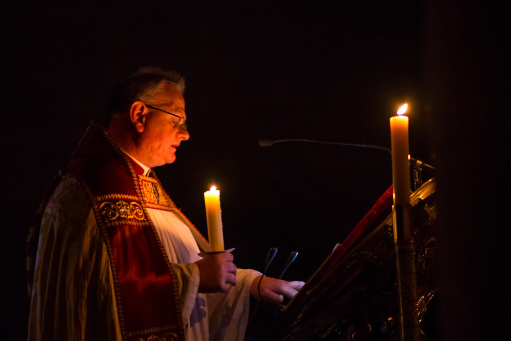 A priest is lit only by the candle he is holding, and a candle that is lighting up the Bible he reads from