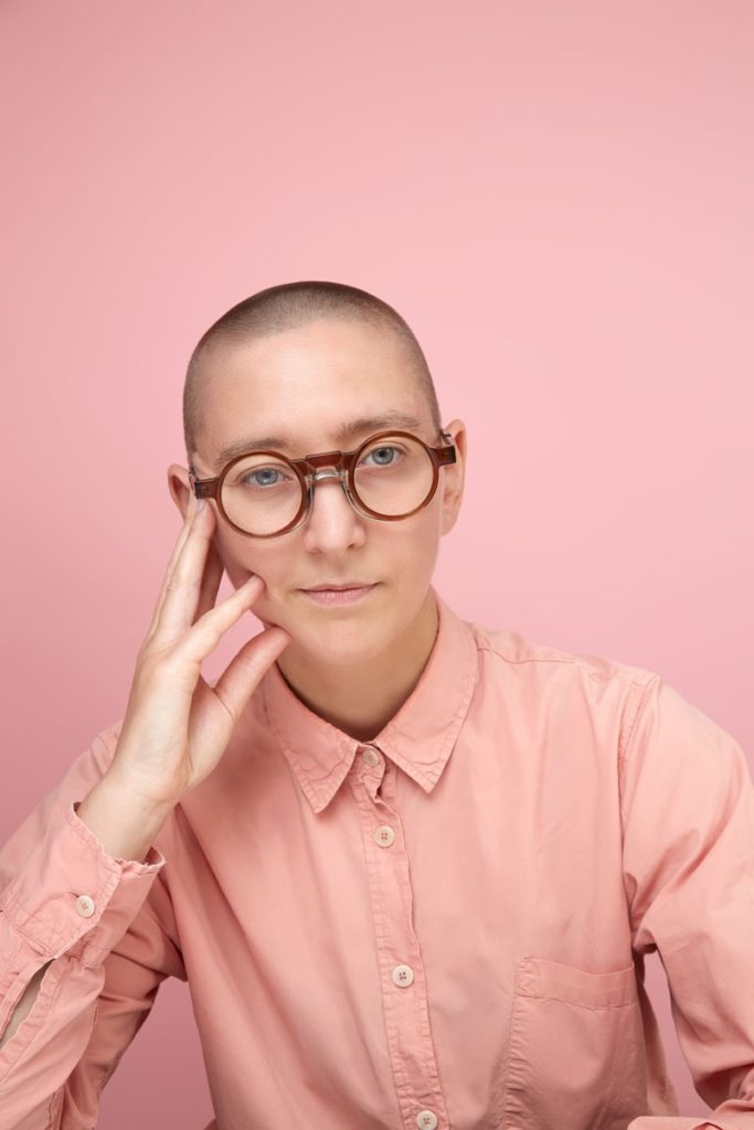 Headshot of Amy Bell. Amy is wearing a pale pink buttoned up shirt and glasses, and is leaning gently against the fingers of her hand which is resting on her face.