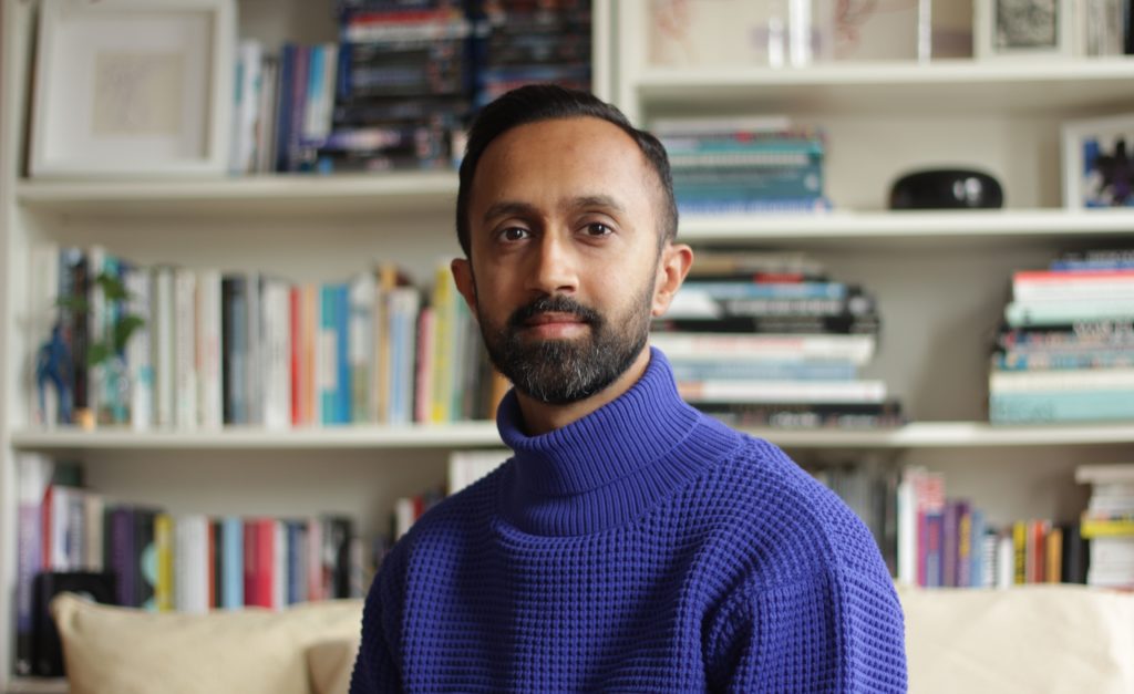 A headshot of Hetain Patel. He is wearing a rich purple roll neck jumper, and is looking into the camera. Behind him is an out of focus book shelf.