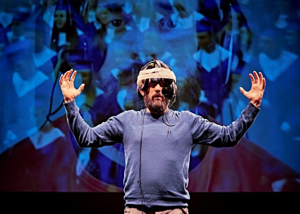 Johnny is standing on stage with a blue abstract projection lighting up the set behind him. He has a homemade contraption on his head which is made from gaffa tape, headphones and wires. His arms are reaching out either side of him.