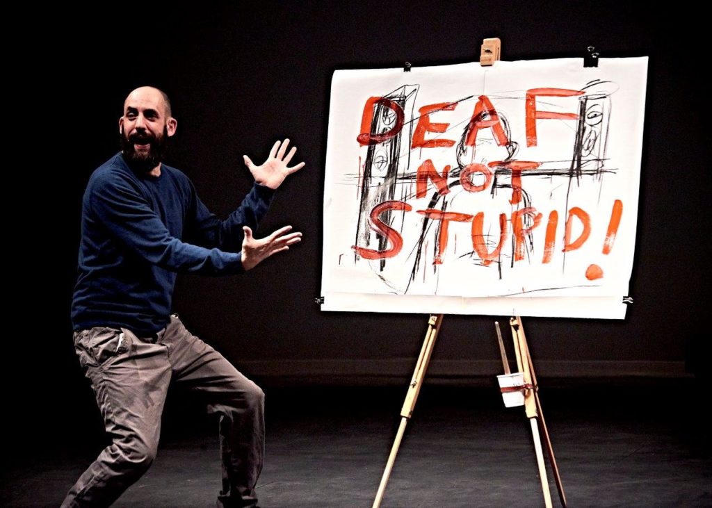 Johnny is standing on stage next to a large easel which holds a piece of paper reading 'Deaf not stupid' in red capital letters.