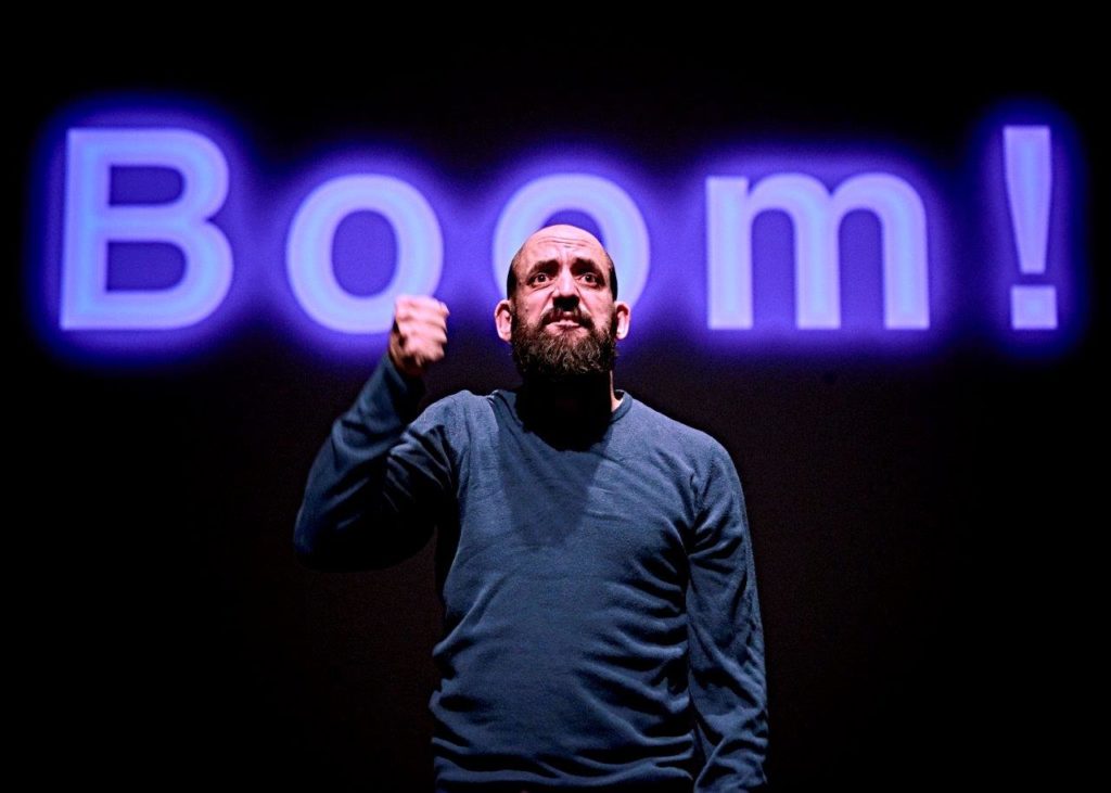 Johnny is standing on stage in front of a projection of the word 'Boom!' in bright purple letters.