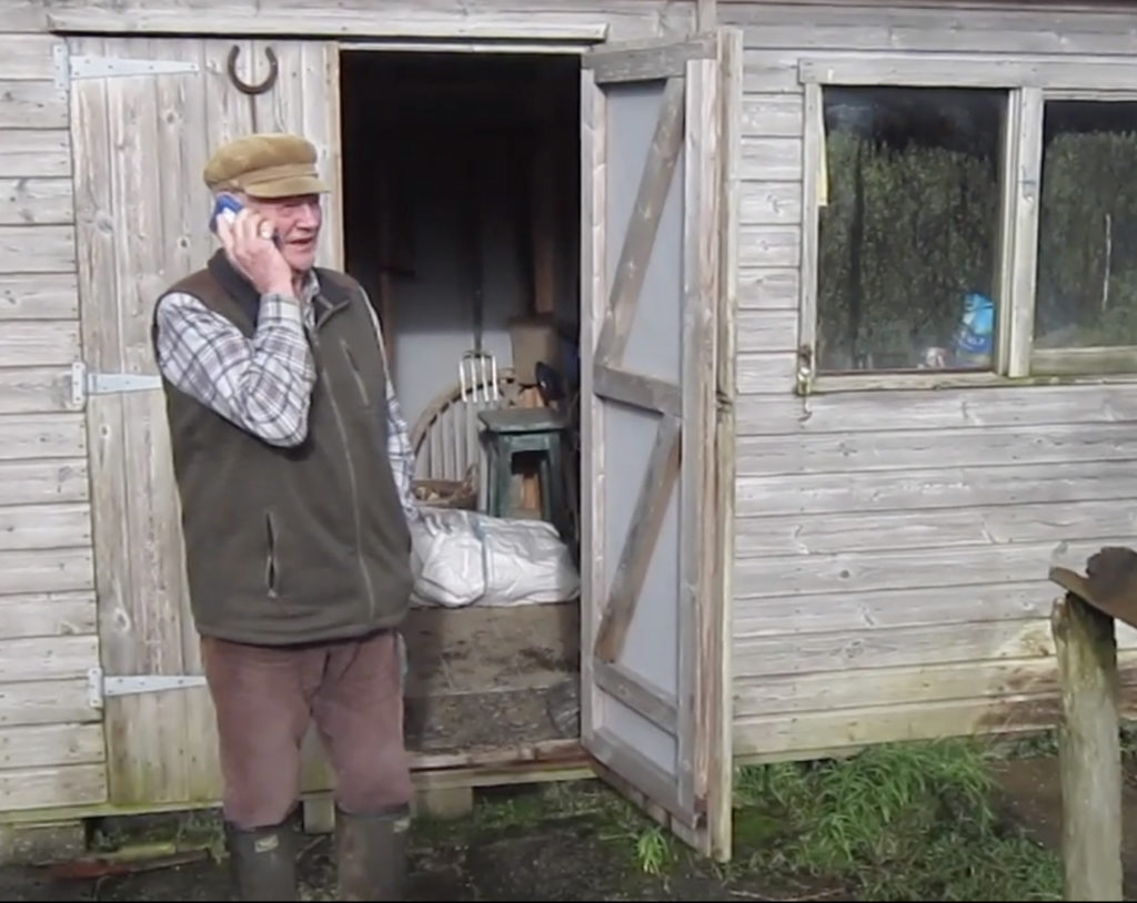 A man wearing a khaki fleece and brown trousers tucked into green wellies is standing outside a brown wooden shed. He is holding a phone to his ear.