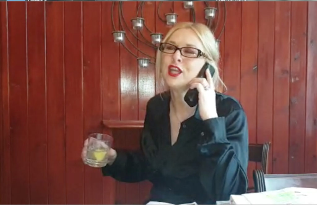 A woman is speaking on the phone which she is holding up to her ear. She is holding a drink in her other hand, which looks like a gin and tonic. She is wearing black glasses, a smart black button up shirt, and red lipstick.