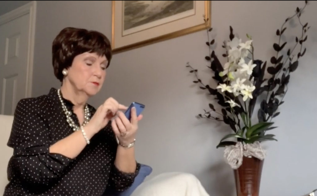 A woman is sitting on a cream armchair in a living room with pale blue walls. She is wearing a black top and a large silver necklace, and it holding a phone in her hand. She is pursing her red lips.