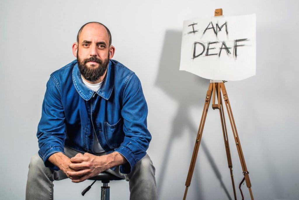 Johnny is sitting on a chair, looking straight towards the camera. He is wearing a blue jacket and grey jeans. He is sitting next to an easel which holds a piece of paper. On the piece of paper is written 'I am deaf' in capital letters.