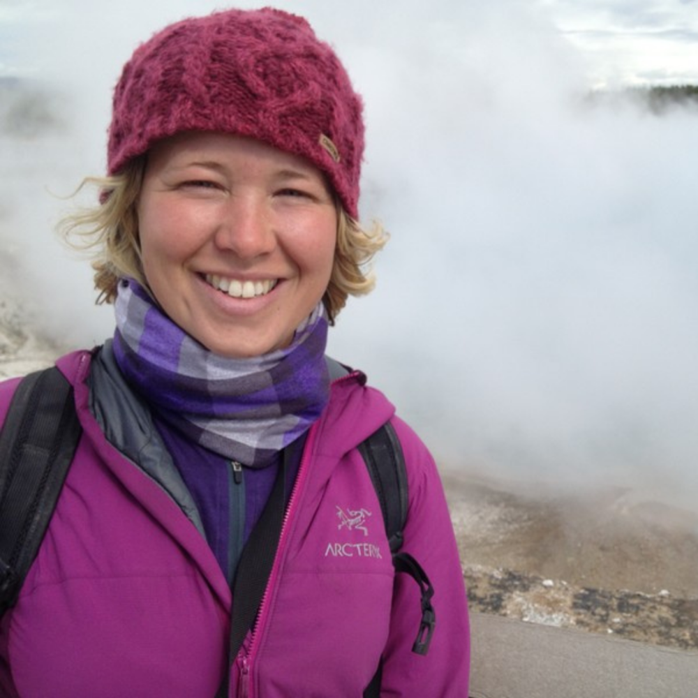 A white woman with short blonde hair is smiling at the camera. She is wearing a purple wooly hat, a checked purple scarf, and a purple coat. The background behind her is obscured by mist.
