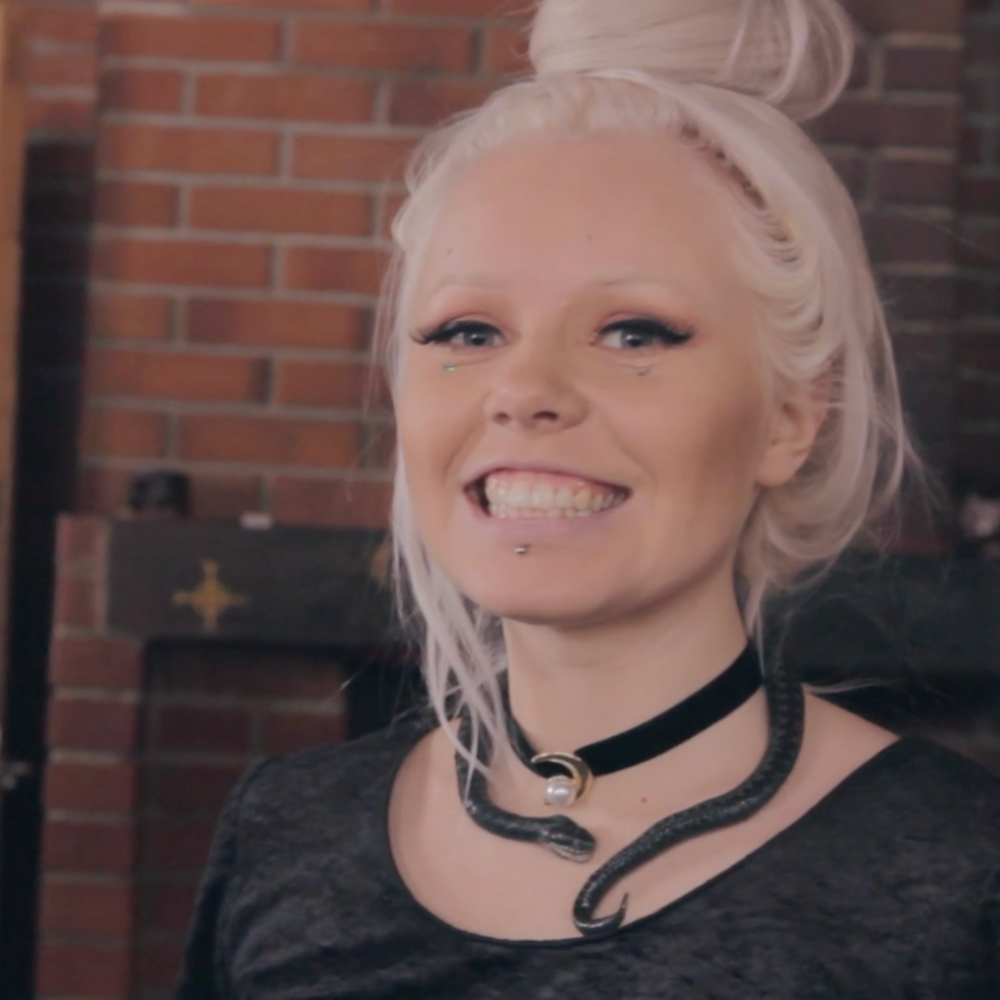 A white woman with white blonde hair tied into a messy bun is smiling at the camera. She has winged black eyeliner, long black eyelashed and silver sparkles dotted under her eyes. She has a snaked wrapped around her neck like a necklace.