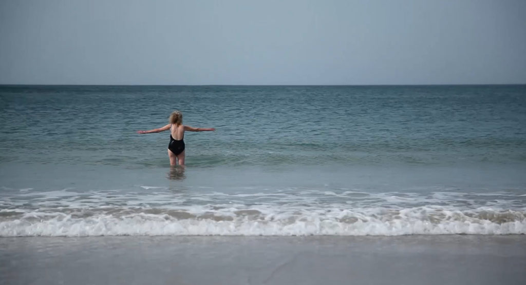 A woman is standing in the sea with her arms stretched out either side of her. It's a sunny day and the water is calm.