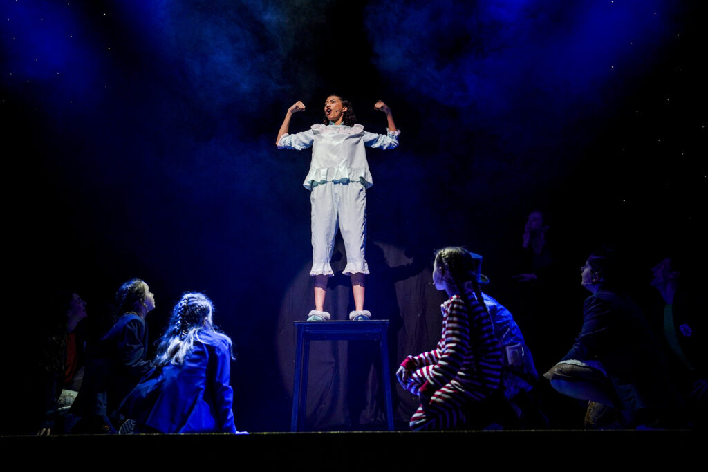 A woman dressed in blue pyjamas stands on the stage with her arms in a strong man position. Other children sit at her feet. 